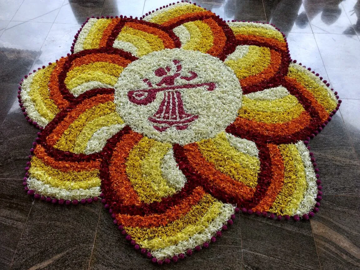 The Onam floral carpet laid for the 2017 celebration — concentric petal rings in white, gold and orange around a central medallion bearing the trust’s veena emblem.