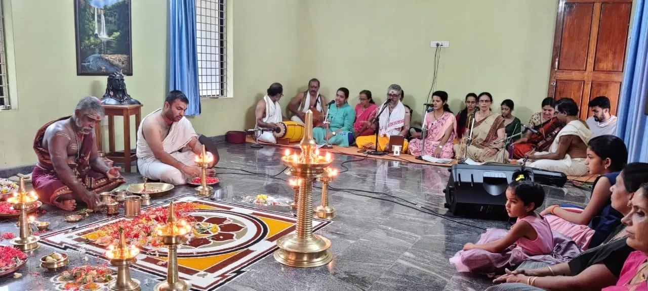A wide view of the Narayaneeyam hall during the second Srividya Pooja: the priests at a lamp-lit Srichakra kolam, with families seated in rows along the walls.
