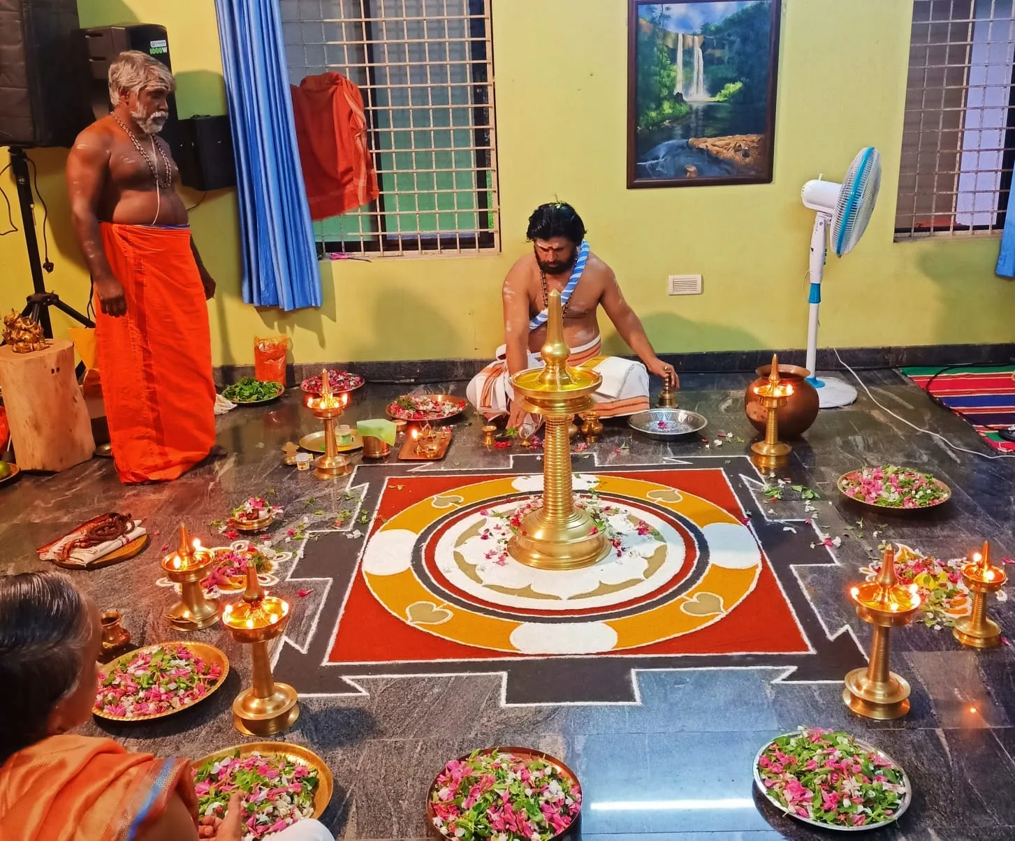 One priest stands while another sits in worship before an elaborate coloured Srichakra kolam, ringed with brass lamps, flowers and offerings.