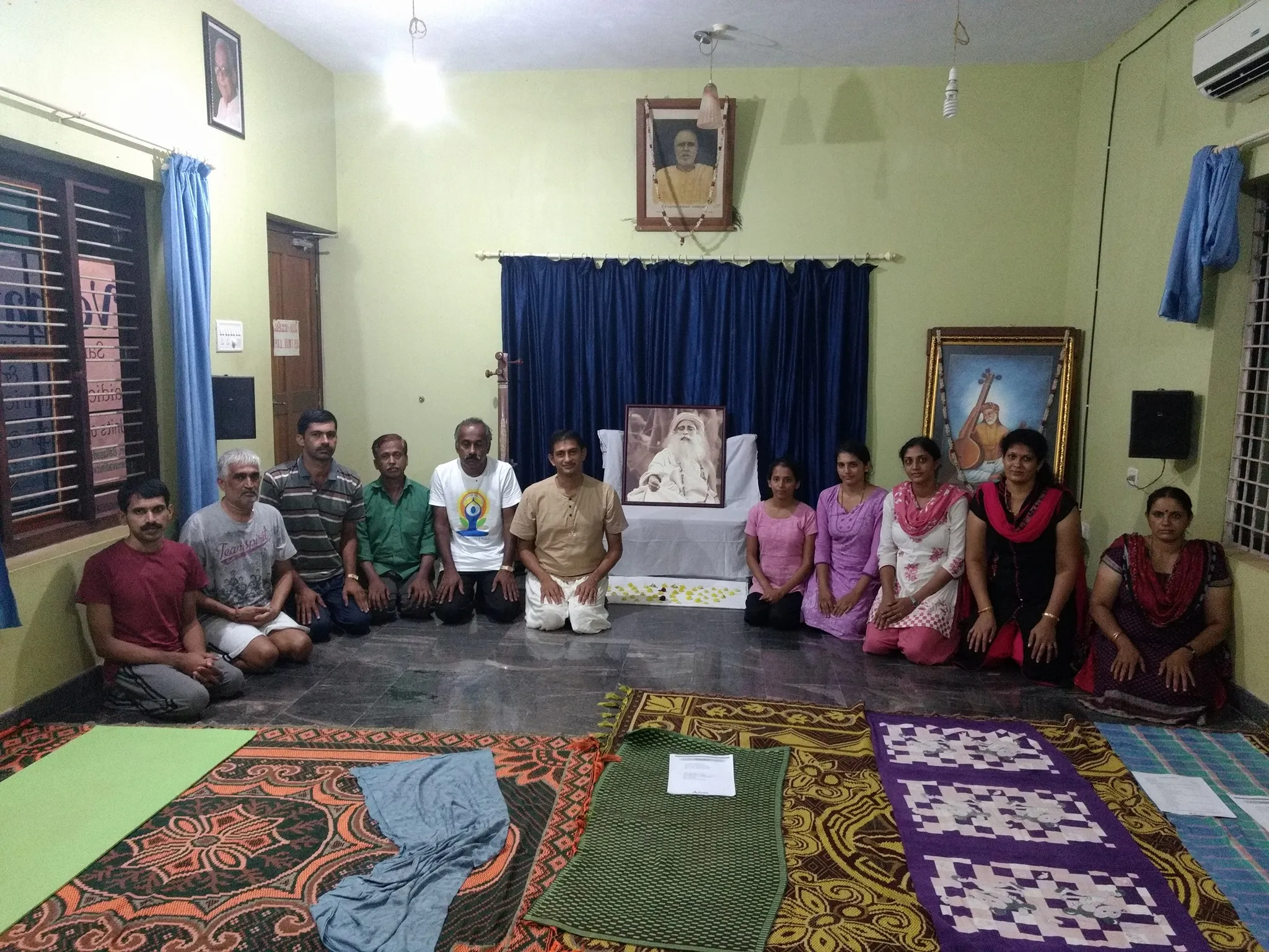 Participants of the Surya Kriya programme seated on their mats in a wide semicircle in the Narayaneeyam hall, the teacher at the centre before a framed portrait.
