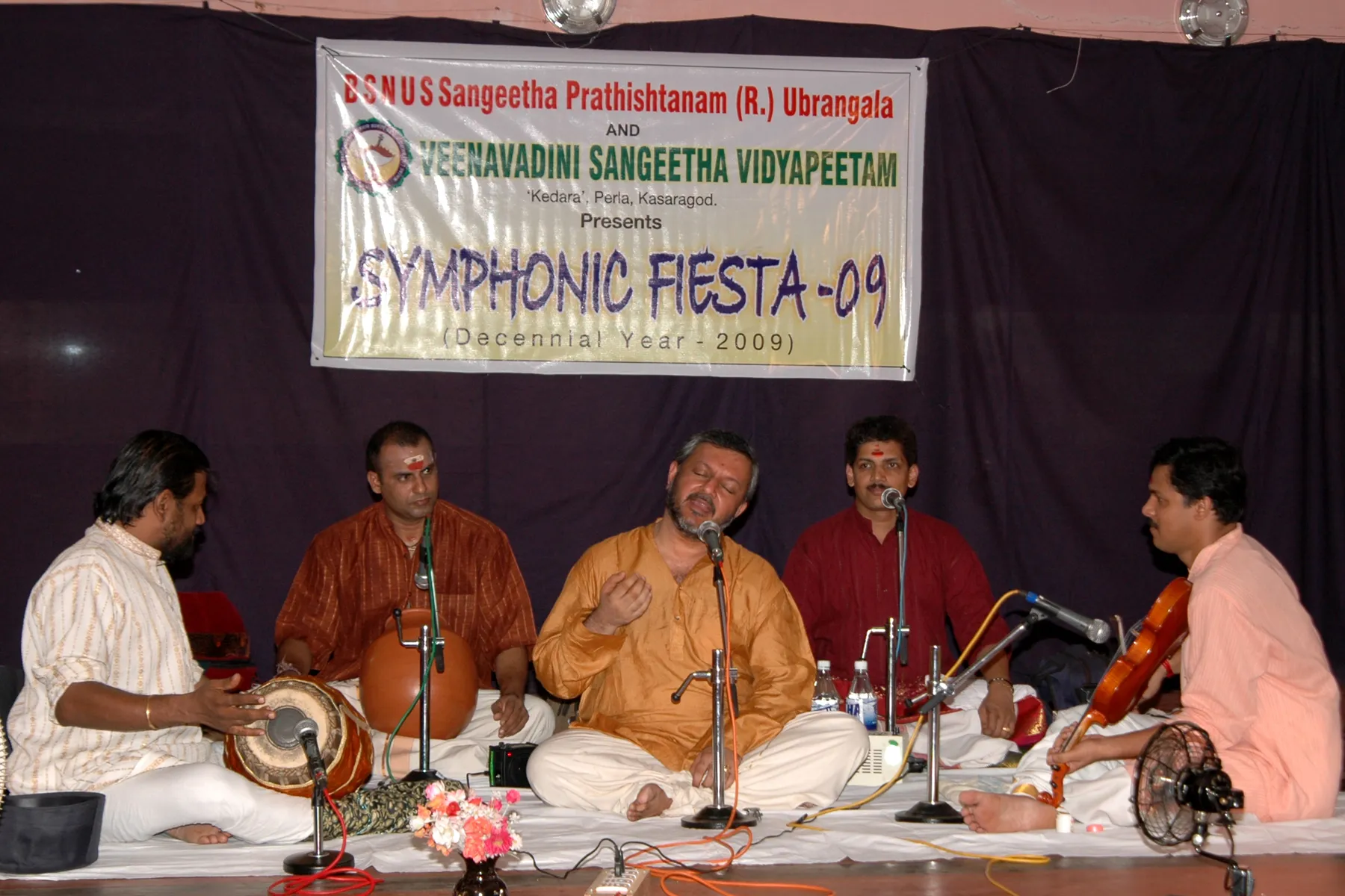 The Symphonic Fiesta 2009 concert in progress, the ensemble seated in a row beneath the festival banner on a maroon-draped stage.