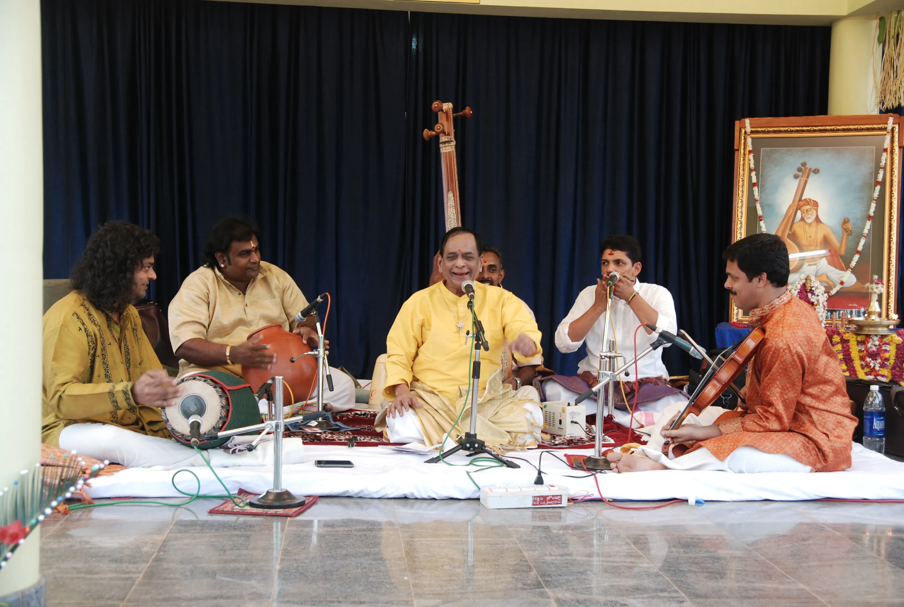 Padmavibhushan Dr. M. Balamuralikrishna singing at Narayaneeyam in 2011, seated in yellow at the centre of his accompanists with a painting of Saraswathi behind the stage.