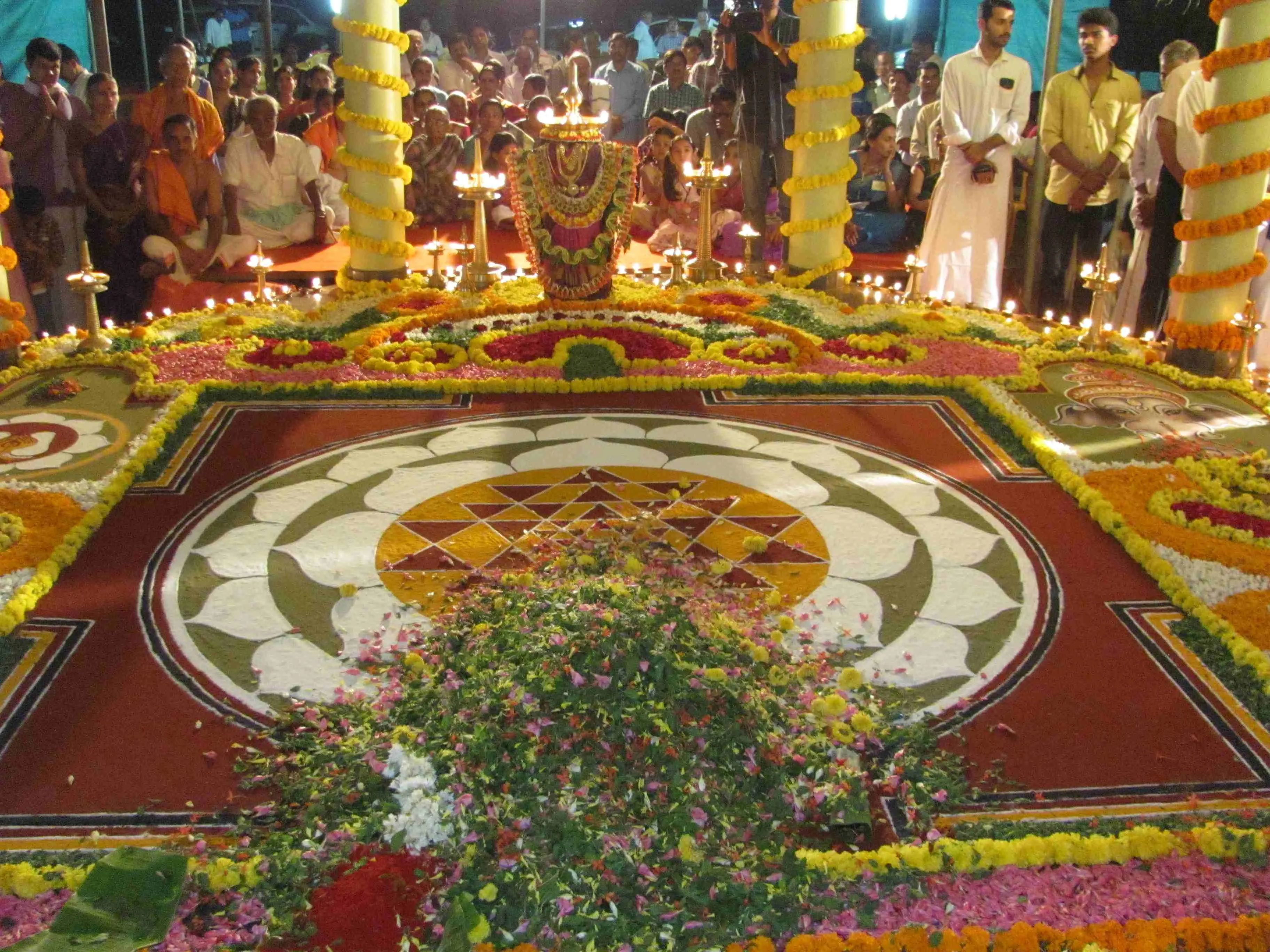 A large mandala of coloured flower petals spread across the hall floor for Veda Nada Tharangini 2016, ringed by tall lamps and by the standing gathering.