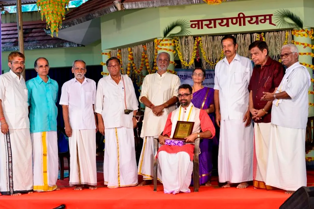 A felicitation on the Narayaneeyam stage during Veda Nada Yoga Tharangini 2017, the honoured guest seated with a plaque while trustees and well-wishers stand around him.