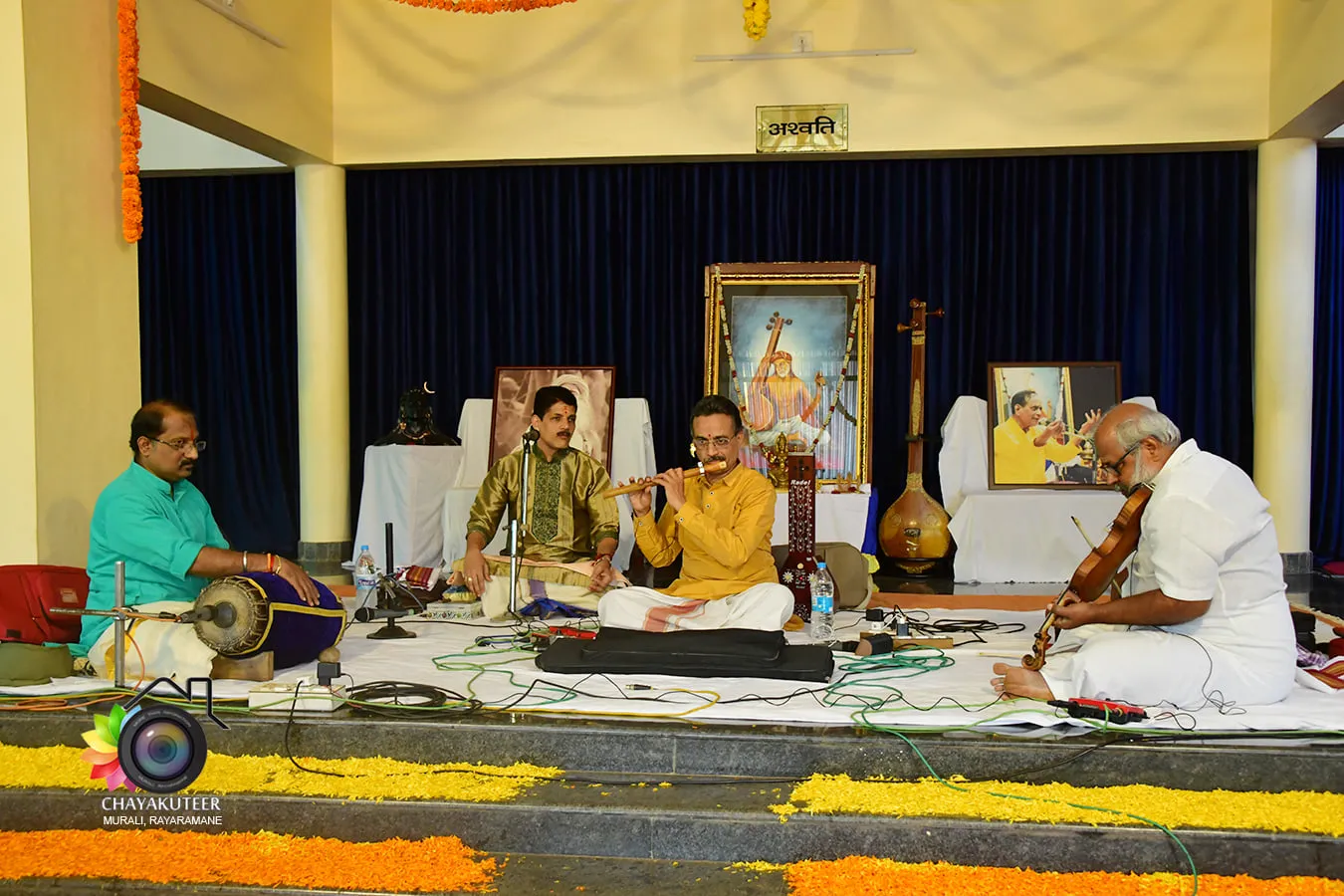 Musicians seated on the Narayaneeyam stage during Veda Nada Yoga Tharangini 2021 — mridangam, vocal, tambura and violin — framed by garlanded portraits behind them.