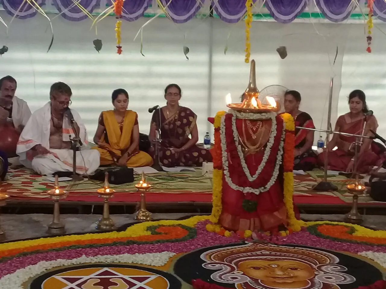 Veenavadini’s singers seated on either side of a garlanded deity at Alappuzha, singing the Navavarana Krithis over a coloured floral kolam.